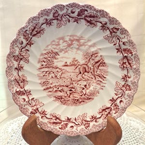 May include: A white ceramic plate with a pink and white floral design. The plate features a hunting scene with a rider on horseback and hounds chasing a fox.