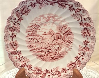 May include: A white ceramic plate with a pink and white floral design. The plate features a hunting scene with a rider on horseback and hounds chasing a fox.