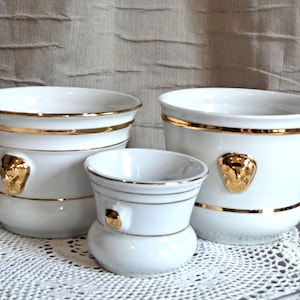 May include: Three white ceramic plant pots with gold trim and a gold Medusa head emblem. The pots are different sizes, with a small, medium, and large pot. The pots are on a white lace doily.