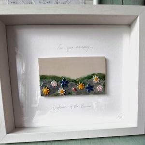 Può includere: Un'immagine incorniciata bianca con una piastrella in ceramica che raffigura una collina verde con fiori colorati e il testo "Per il tuo ricordo... Fiori selvatici del Barrow"