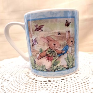 Könnte beinhalten: Eine blaue Keramiktasse mit einem Bild von zwei Kaninchen aus der Peter Rabbit-Geschichte. Die Kaninchen befinden sich in einer Gartenlandschaft mit Vögeln und Schmetterlingen. Die Tasse hat ein blaues und weißes Blumenmuster am Rand.