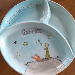 Peut inclure: Une assiette divisée bleu clair avec une illustration peinte à la main du Petit Prince. L'assiette porte les mots "Notre petit prince" et le nom "Gabriel". La décoration comprend des étoiles, une planète et un renard.