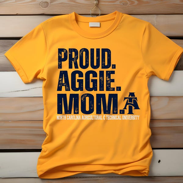 Może przedstawiać: Musztardowa żółta koszulka z granatowym napisem "PROUD. AGGIE. MOM." i logo North Carolina Agricultural & Technical University. Koszulka jest prezentowana na drewnianym tle.