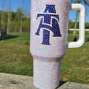 May include: A white stainless steel tumbler with a blue and white rhinestone design. The tumbler has a white handle and a clear lid.