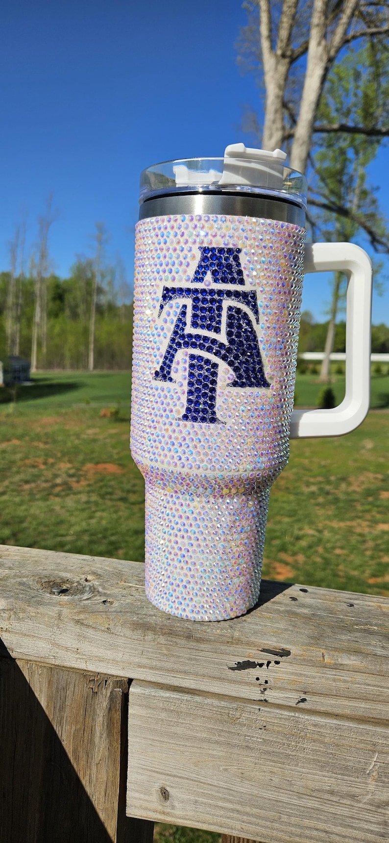 May include: A white stainless steel tumbler with a blue and white rhinestone design. The tumbler has a white handle and a clear lid.