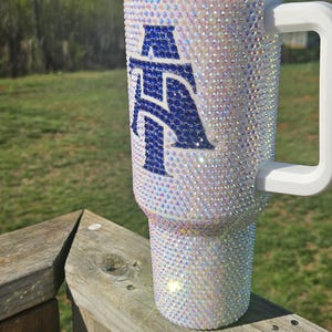 May include: A white tumbler with a blue letter "A" covered in iridescent rhinestones. The tumbler has a white lid and a white handle.