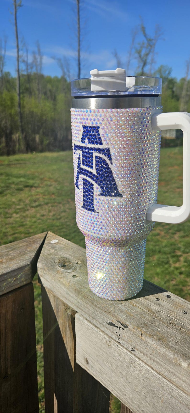 May include: A white tumbler with a blue letter "A" covered in iridescent rhinestones. The tumbler has a white lid and a white handle.