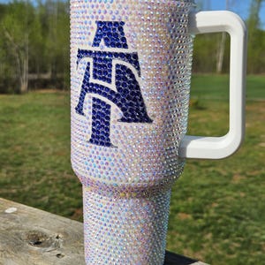 May include: A white tumbler with a blue and white letter "A" design covered in iridescent rhinestones. The tumbler has a white handle and a clear lid.