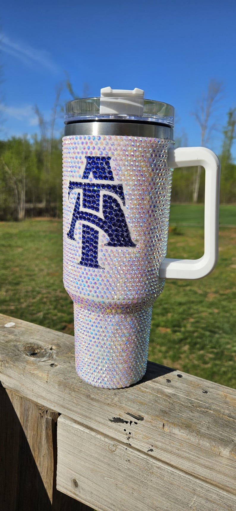 May include: A white tumbler with a blue and white letter "A" design covered in iridescent rhinestones. The tumbler has a white handle and a clear lid.