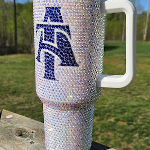 May include: A white stainless steel travel mug with a blue and white letter "A" design. The mug has a white handle and a clear lid. The mug is covered in iridescent rhinestones.
