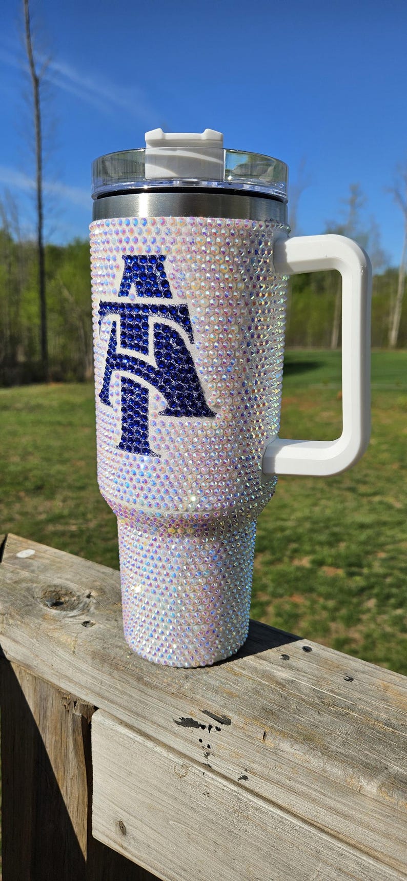May include: A white stainless steel travel mug with a blue and white letter "A" design. The mug has a white handle and a clear lid. The mug is covered in iridescent rhinestones.