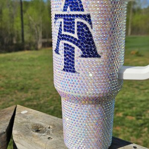 May include: A white tumbler with a blue and white letter "A" design. The tumbler is covered in iridescent rhinestones.