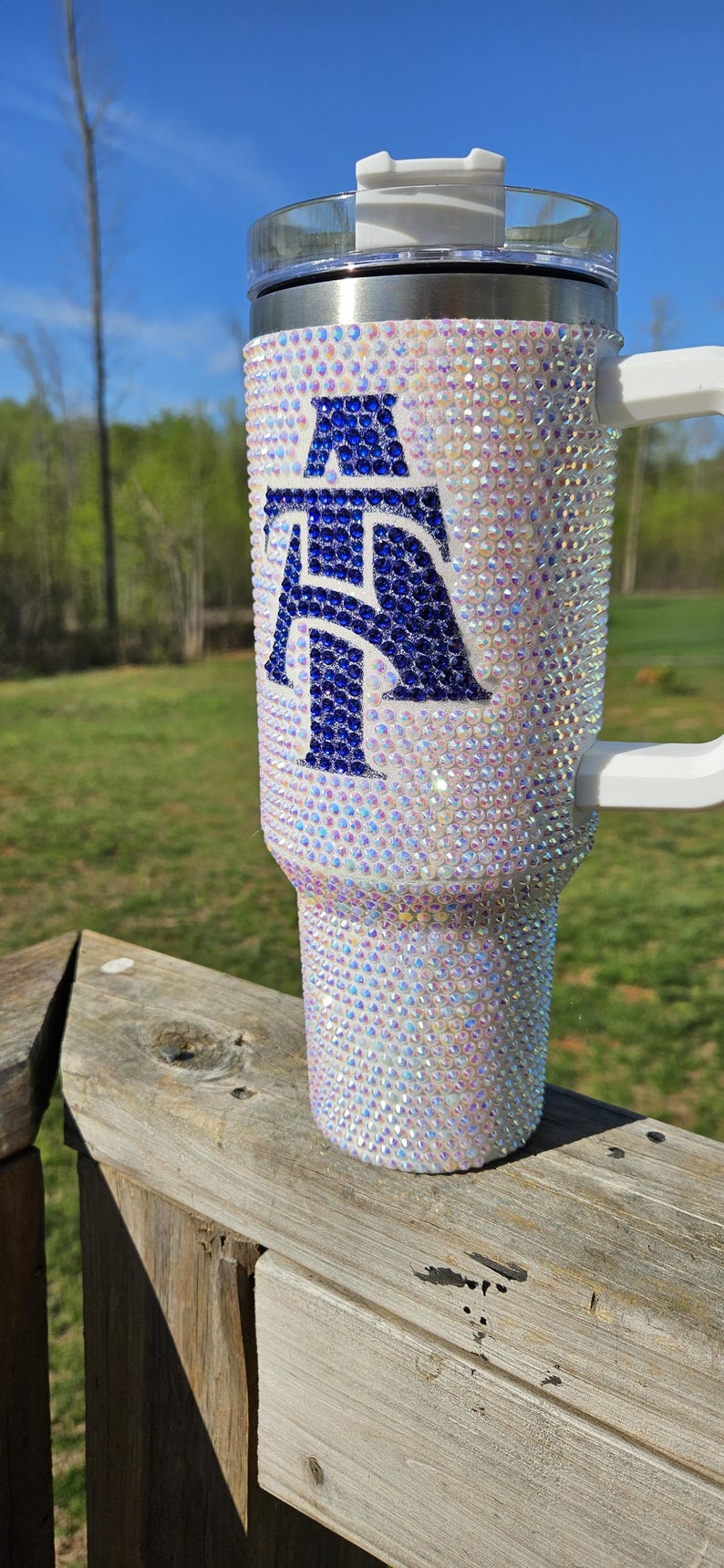 May include: A white tumbler with a blue and white letter "A" design. The tumbler is covered in iridescent rhinestones.