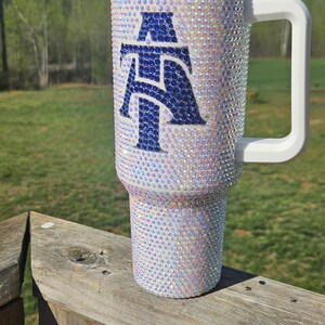 May include: White stainless steel tumbler with a blue and white letter "A" design covered in iridescent rhinestones. The tumbler has a white lid and a white handle.
