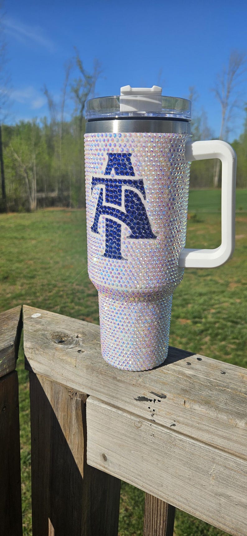 May include: White stainless steel tumbler with a blue and white letter "A" design covered in iridescent rhinestones. The tumbler has a white lid and a white handle.