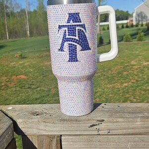 May include: A white tumbler with a blue and white letter "A" design. The tumbler is covered in iridescent rhinestones and has a white lid and handle.