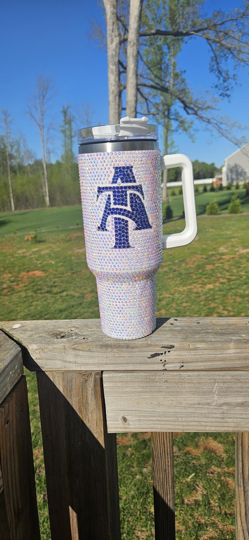 May include: A white tumbler with a blue and white letter "A" design. The tumbler is covered in iridescent rhinestones and has a white lid and handle.