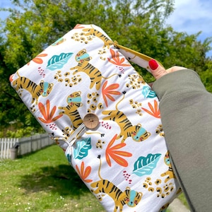 Könnte beinhalten: Eine weiße, rechteckige Wickelauflage mit buntem Tigerdruck. Das Design zeigt orange, türkisfarbene und gelbe Blätter und Tiger mit blauen Gesichtsmasken. Ein Holzknopf und ein elastischer Verschluss sichern die Auflage.