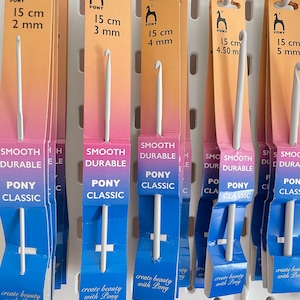 Könnte beinhalten: Nahaufnahme einer Auswahl weißer Häkelnadeln, die mit dem Markennamen "Pony" und der Größe in Zentimetern und Millimetern gekennzeichnet sind. Die Nadeln werden auf einer weißen Lochwand präsentiert.