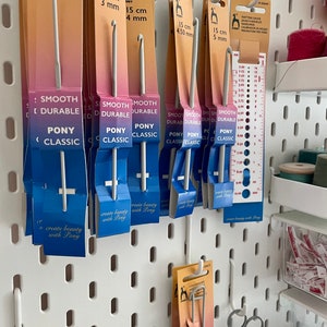 May include: A white pegboard with various sizes of Pony brand crochet hooks hanging on it. The hooks are labeled with their size in centimeters and millimeters. The pegboard also has a ruler and a small container of yarn scraps.