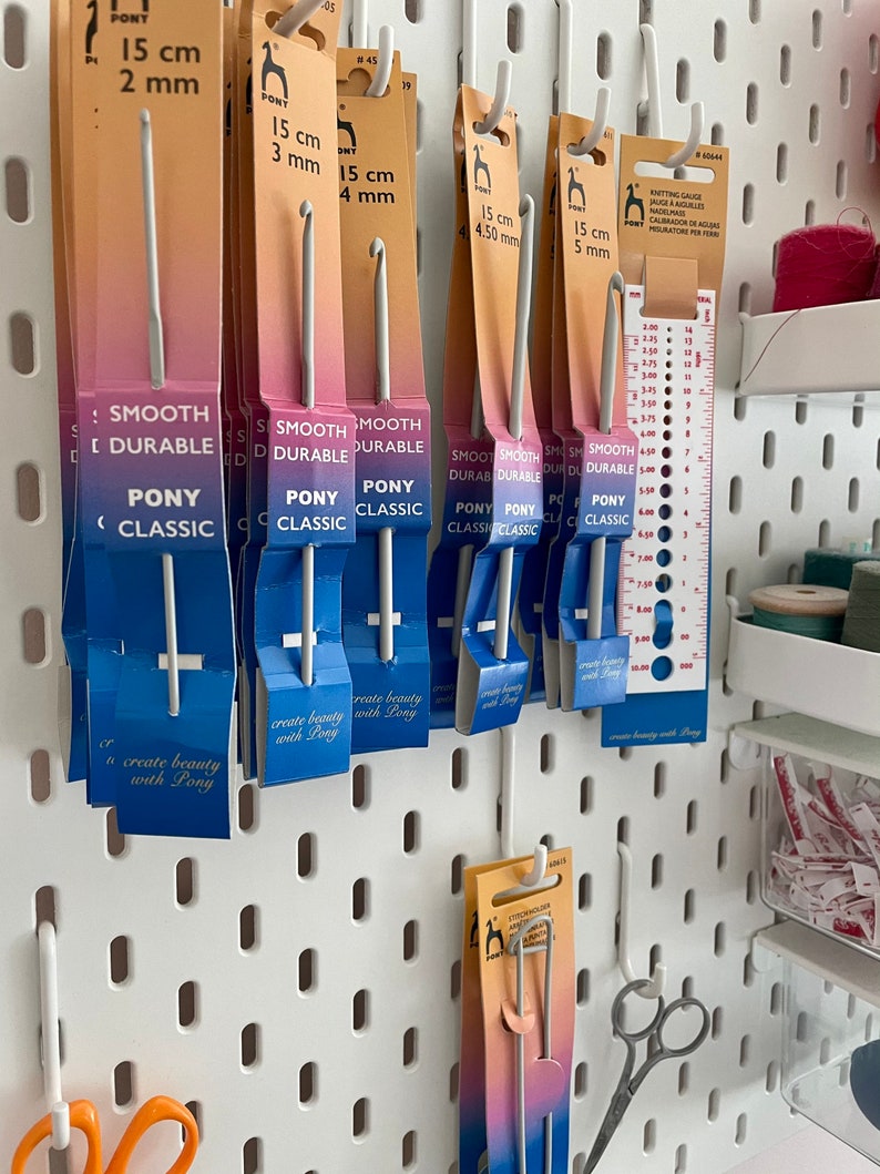 May include: A white pegboard with various sizes of Pony brand crochet hooks hanging on it. The hooks are labeled with their size in centimeters and millimeters. The pegboard also has a ruler and a small container of yarn scraps.