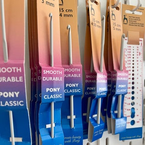 May include: A close-up of a set of Pony Classic crochet hooks in various sizes. The hooks are smooth and durable, and they are packaged in blue and white packaging with the Pony Classic logo. The packaging also includes the size of each hook in centimeters and millimeters.