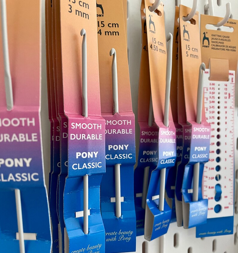 May include: A close-up of a set of Pony Classic crochet hooks in various sizes. The hooks are smooth and durable, and they are packaged in blue and white packaging with the Pony Classic logo. The packaging also includes the size of each hook in centimeters and millimeters.