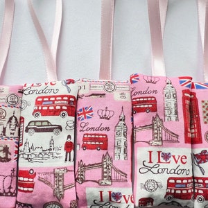 以下が含まれることがあります： ロンドンをテーマにしたピンクの布製のサシェが5つ。各サシェは、ビッグベン、タワーブリッジ、2階建てバスなど、ロンドンの象徴的なランドマークを異なるデザインで特徴としています。サシェはピンクのリボンで結ばれています。
