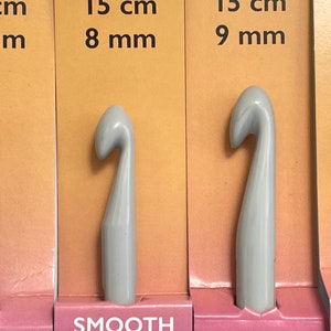 May include: Four gray crochet hooks with smooth finishes. The hooks are labeled with their sizes in centimeters and millimeters: 15 cm 7 mm, 15 cm 8 mm, 15 cm 9 mm, and 10 mm. The hooks are displayed on a pink and orange background.