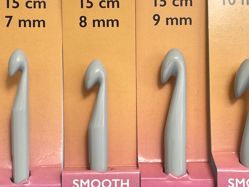 May include: Four gray crochet hooks with smooth finishes. The hooks are labeled with their sizes in centimeters and millimeters: 15 cm 7 mm, 15 cm 8 mm, 15 cm 9 mm, and 10 mm. The hooks are displayed on a pink and orange background.