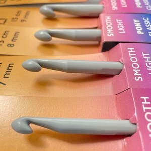 May include: A set of gray crochet hooks with different sizes labeled in millimeters. The hooks are arranged in a row on a colorful background with the words "Smooth", "Light", "Pony", and "Classic" printed on the labels.