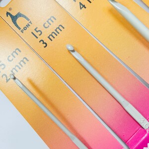 May include: Three Pony brand crochet hooks in different sizes. The hooks are 3 mm, 4 mm, and 5 mm. The hooks are made of a light gray plastic and are packaged on a yellow and pink background.