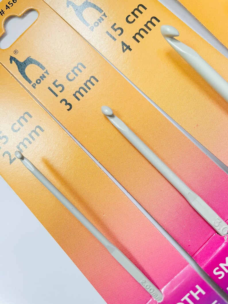 May include: Three Pony brand crochet hooks in different sizes. The hooks are 3 mm, 4 mm, and 5 mm. The hooks are made of a light gray plastic and are packaged on a yellow and pink background.