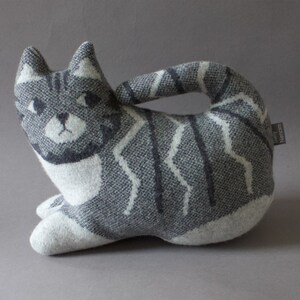 May include: A gray and white striped cat-shaped plush toy with a white face and black eyes. The toy has a label that says "Emily Hencson".