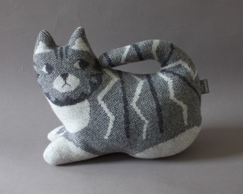 May include: A gray and white striped cat-shaped plush toy with a white face and black eyes. The toy has a label that says "Emily Hencson".