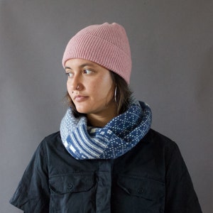 Könnte beinhalten: Eine hellrosa gerippte Strickmütze und ein blau-weiß gemusterter Loop-Schal. Der Schal hat ein geometrisches Design. Die Person trägt ein schwarzes Hemd mit Knöpfen.