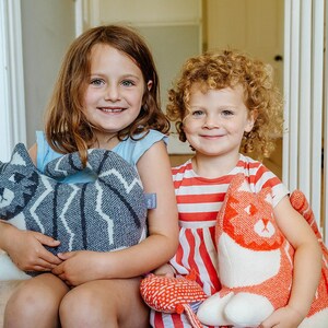 May include: Two young girls are sitting on a wooden floor, each holding a knitted cat toy. The girl on the left is holding a gray and white cat toy, while the girl on the right is holding a red and white cat toy. Both girls are smiling.