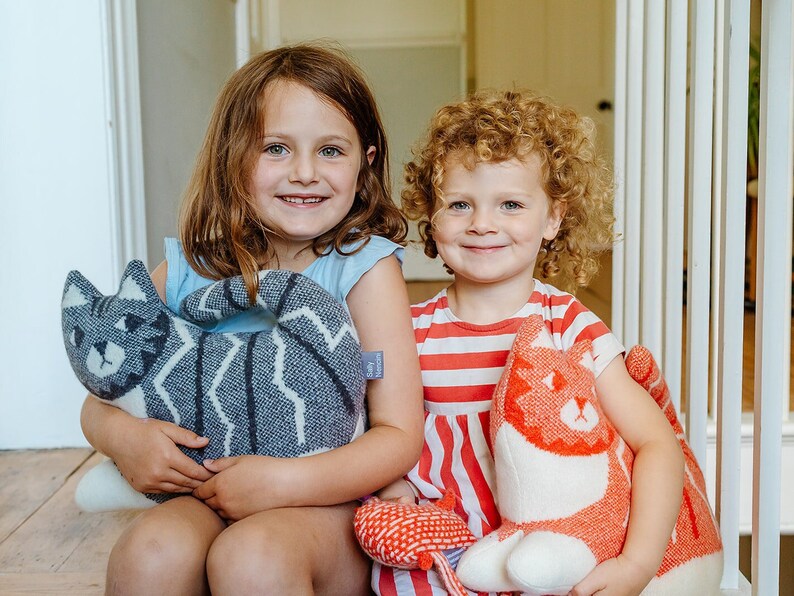 May include: Two young girls are sitting on a wooden floor, each holding a knitted cat toy. The girl on the left is holding a gray and white cat toy, while the girl on the right is holding a red and white cat toy. Both girls are smiling.