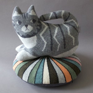 May include: A gray and white striped cat-shaped pillow sits on top of a round, multi-colored striped pillow. The pillows are made of a soft, knitted fabric.