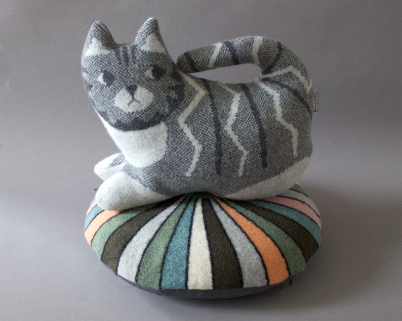 May include: A gray and white striped cat-shaped pillow sits on top of a round, multi-colored striped pillow. The pillows are made of a soft, knitted fabric.