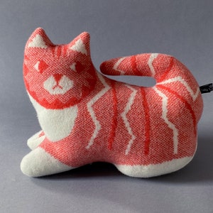 May include: A red and white knitted cat plush toy. The cat is lying on its side with its tail curled around its body. The cat has a white face with red eyes and a pink nose.