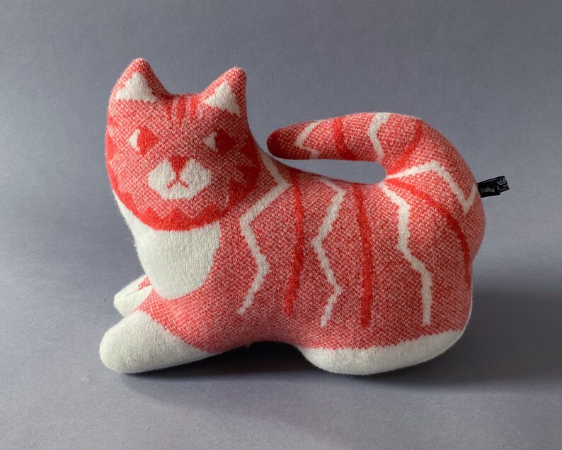 May include: A red and white knitted cat plush toy. The cat is lying on its side with its tail curled around its body. The cat has a white face with red eyes and a pink nose.