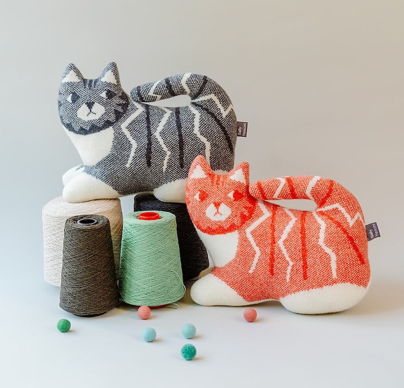May include: Two knitted cat-shaped plush toys, one gray and white, the other orange and white, with yarn spools in the background.