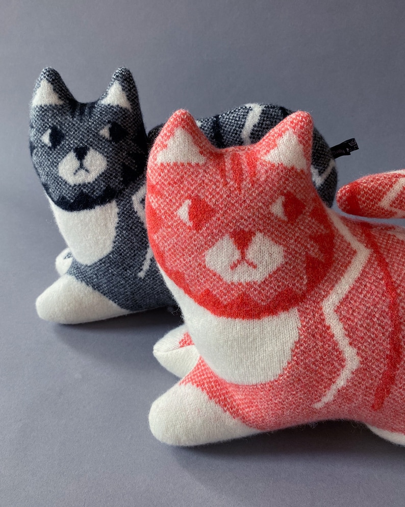 May include: Two knitted cat-shaped plush toys. One is gray and white with black stripes, the other is white with red stripes. Both have black eyes and a white nose.