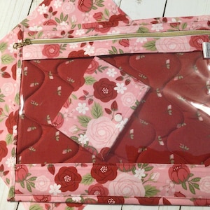 May include: A rectangular zippered pouch featuring a clear vinyl front panel and a pink fabric border decorated with a floral pattern of red and light pink roses with green leaves. The interior lining displays a quilted red rose pattern.