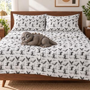 May include: A white bed sheet set featuring a repeating black graphic pattern of a monster in various poses. A plush toy of a sleeping grey monster with textured scales and dorsal fins rests on the center of the bed, which has a wooden headboard.