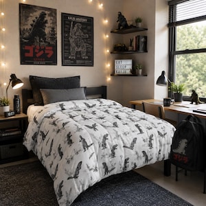 May include: A white comforter featuring a repeating black and grey pattern of Godzilla figures. The bedding is draped over a bed frame with a dark grey pillow and a rectangular bolster pillow. The design includes various poses of the creature.