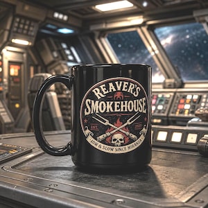 May include: Black ceramic mug featuring the text "Reaver's Smokehouse" in a circular design with crossed meat forks and a skull. The mug also includes the text "Low & Slow Since Miranda" and the year 2017.