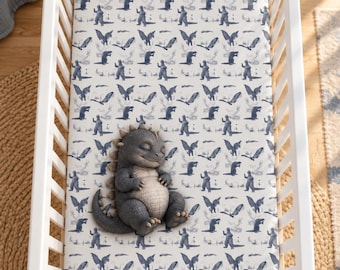 May include: A white crib sheet featuring a repeating blue pattern of various monsters and winged creatures. A plush, gray, textured monster toy with a rounded belly and closed eyes is resting on the center of the sheet inside a white crib.