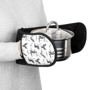 May include: Black and white oven mitts and pot holder set. The mitts have a black quilted exterior and a white interior patterned with gray dragon illustrations. A stainless steel pot with a glass lid is held by the mitts.